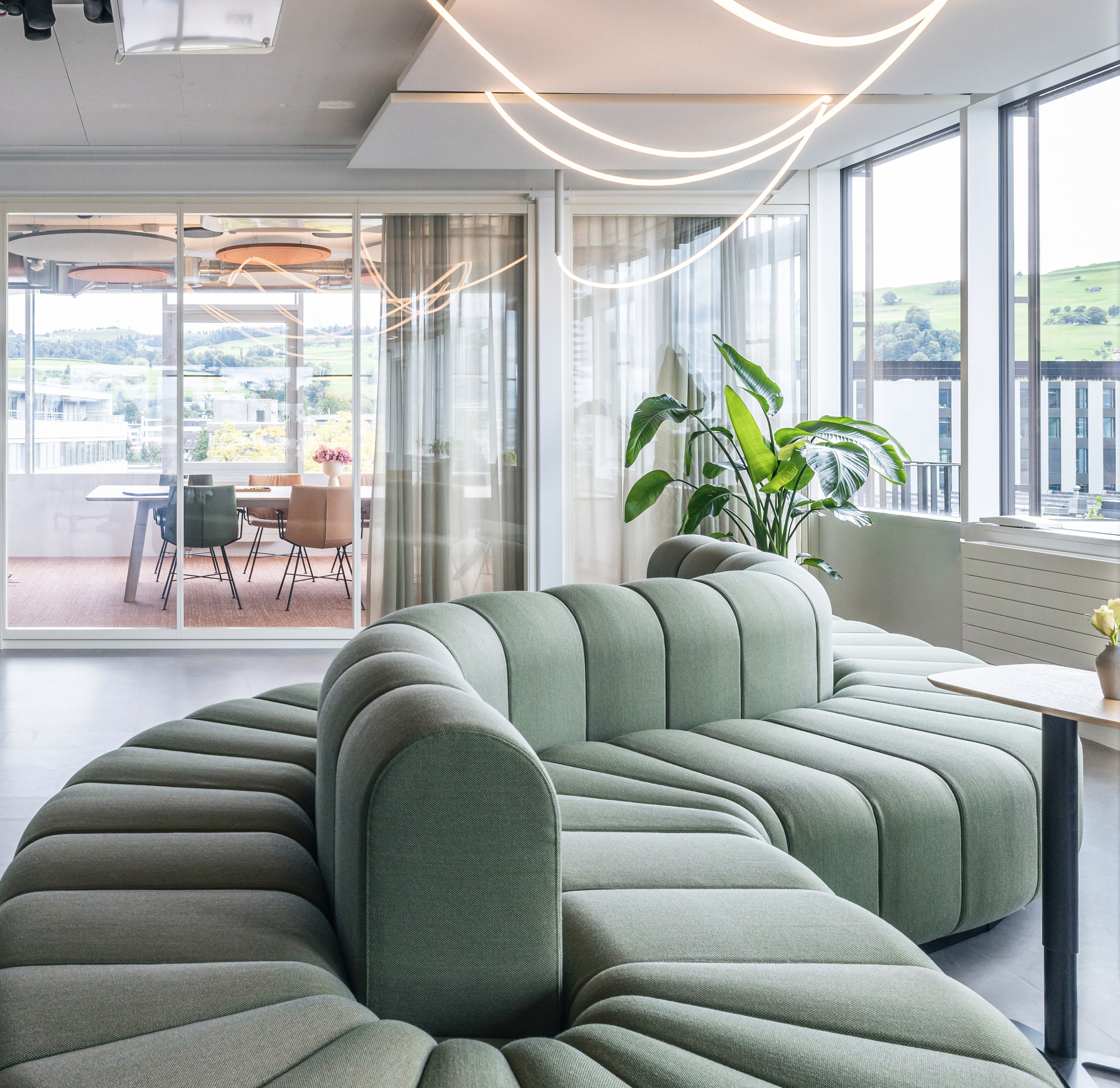 Moderne Bürolounge mit einem geschwungenen, grünen Ecksofa, großen Fenstern, einem verglasten Besprechungsraum und dekorativer Beleuchtung an der Decke.