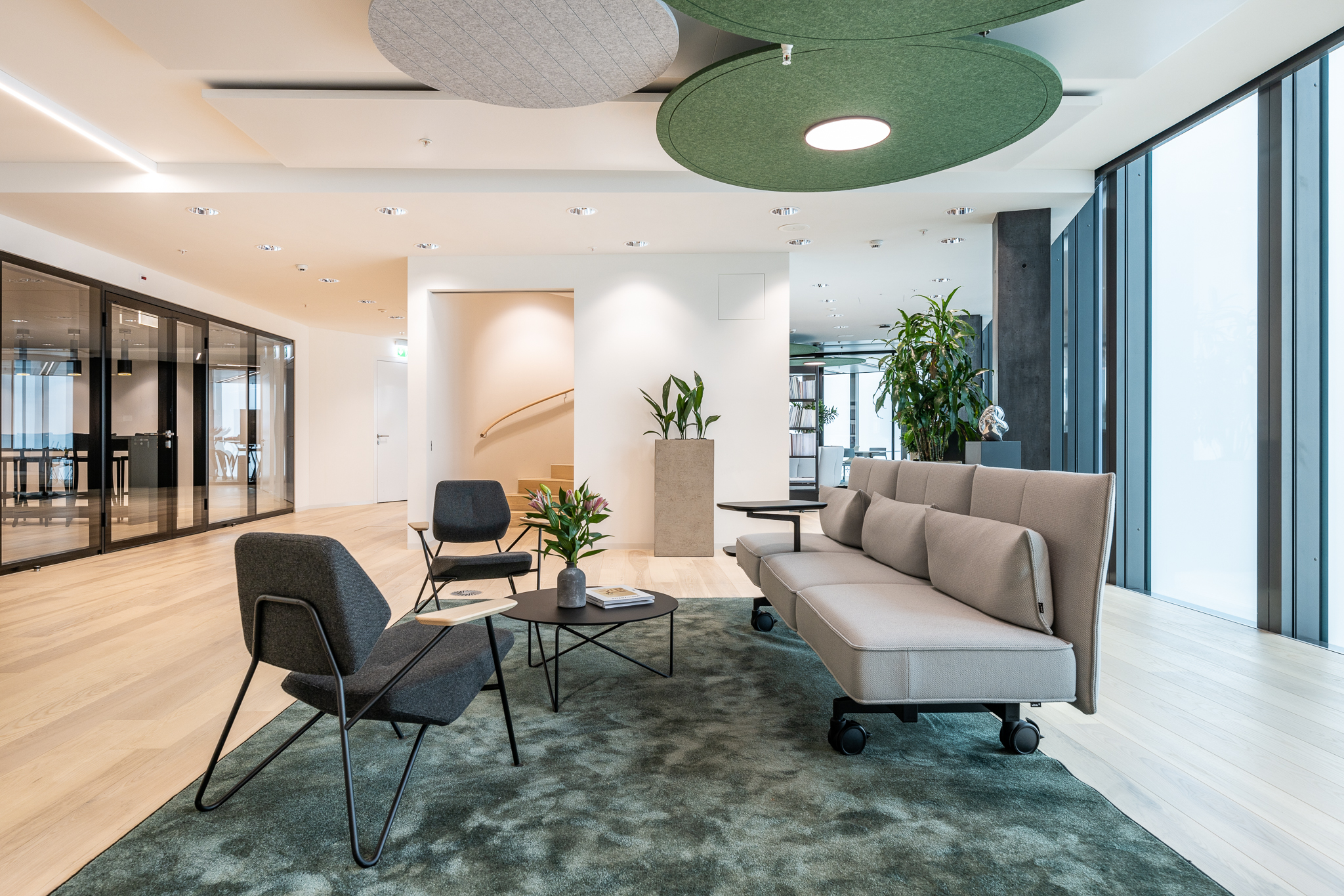 Offene Loungezone mit Sofas und Sesseln auf Teppich, angrenzend an verglaste Meetingräume und helle Arbeitsbereiche.