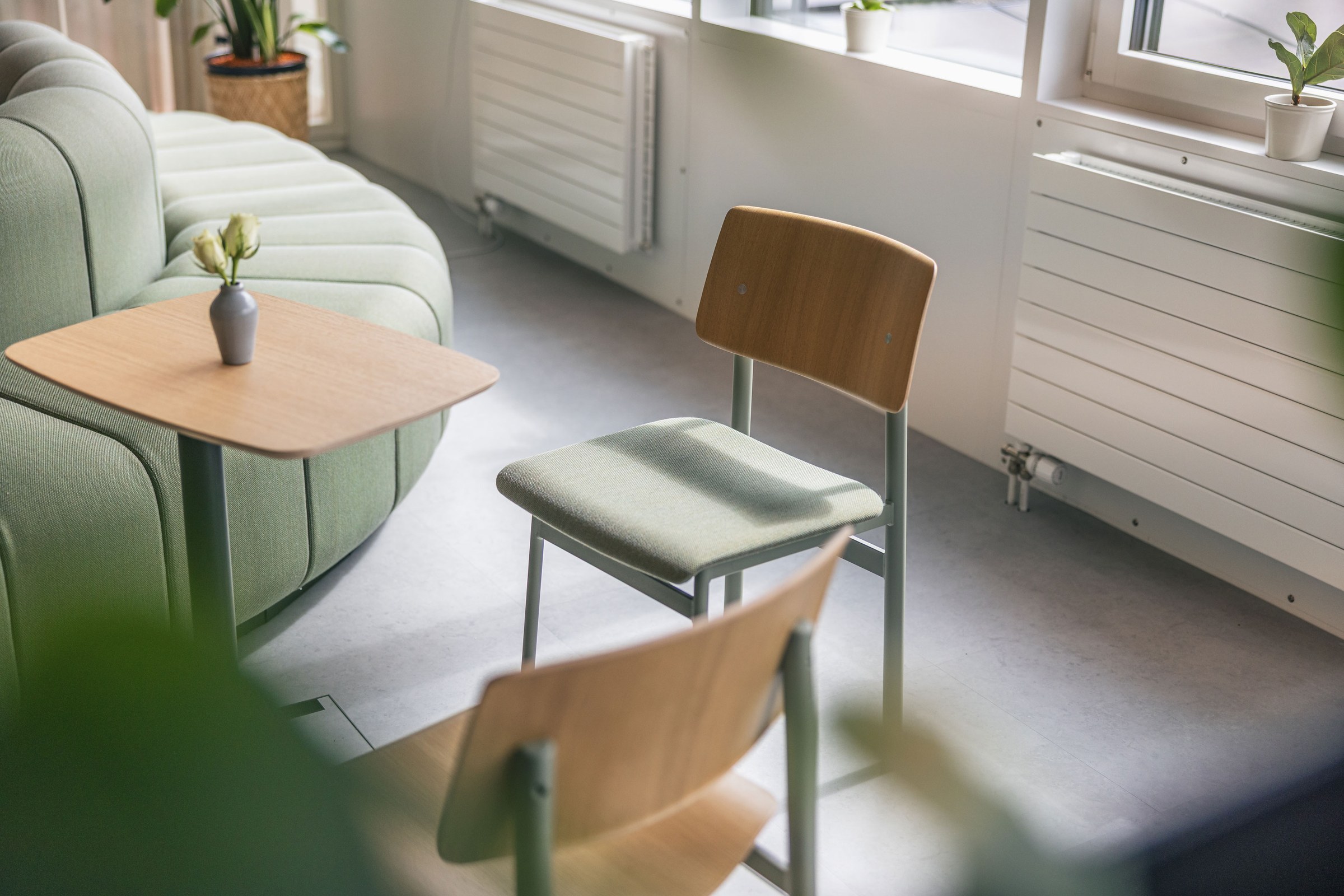 Gemütlicher Büro-Loungebereich mit grünem Sofa, Holzstühlen und kleinen Beistelltischen im Tageslicht.