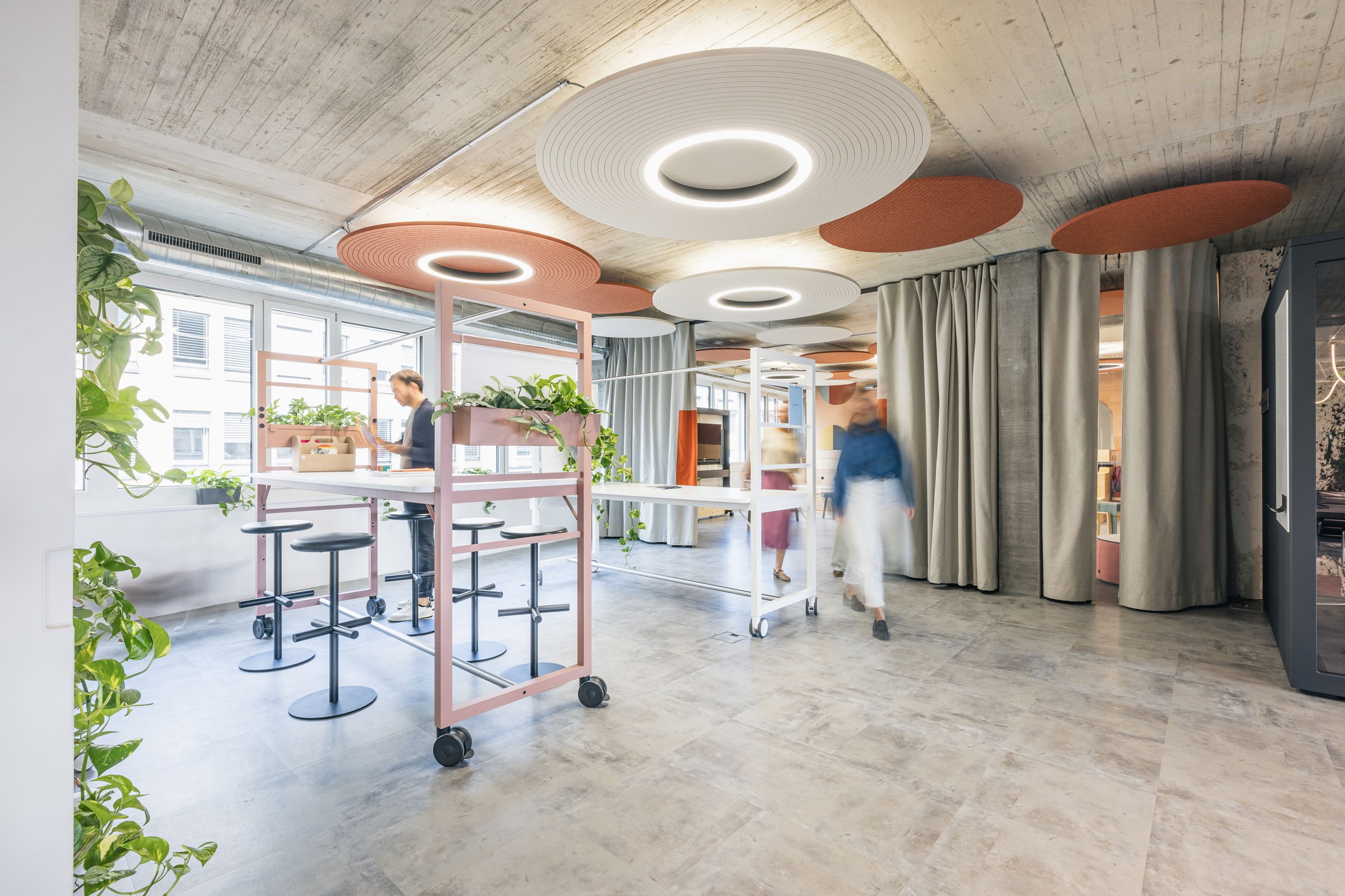 Flexibler Kreativraum mit mobilen Workshopstationen, Pflanzen und runden Akustikdeckenleuchten.