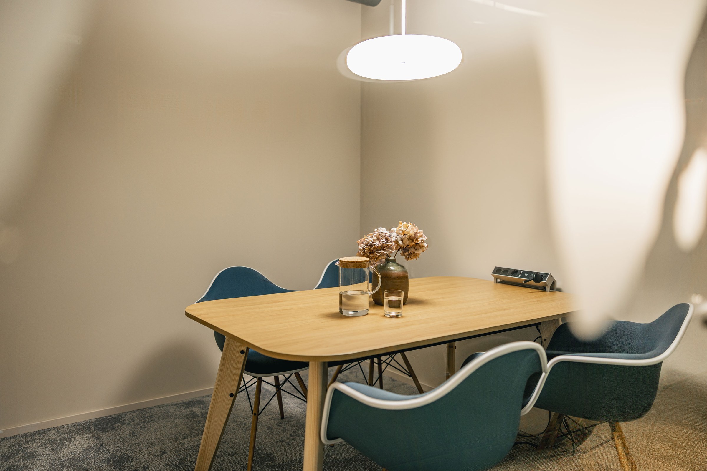 Kleiner Meetingraum mit Holztisch, blauen Stühlen und dekorativer Vase.