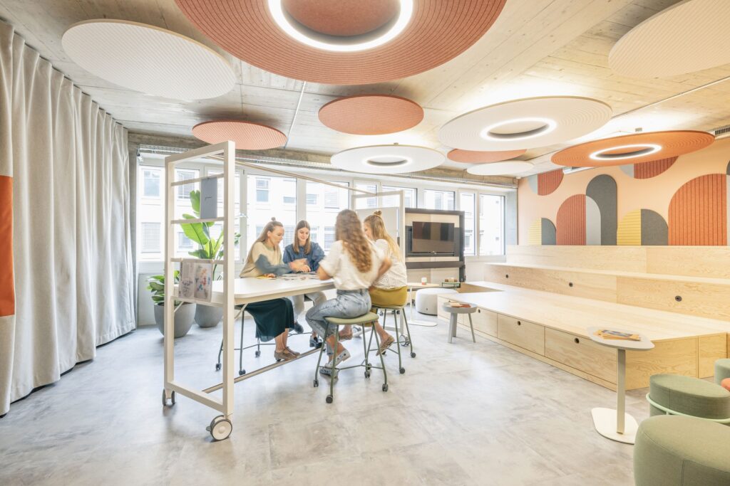 Heller Workshopraum mit runden Akustikdecken, Holzbühne und Teammeeting an einem hohen Arbeitstisch.