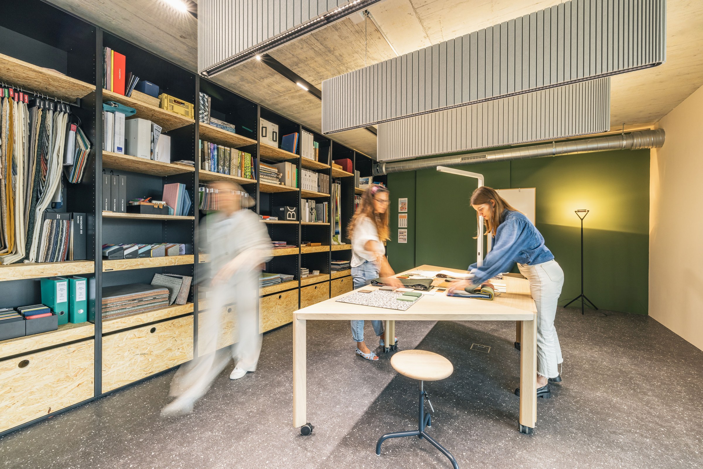 Kreativer Materialraum mit großen Regalen, Stoffmustern und Teamwork an einem zentralen Arbeitstisch.