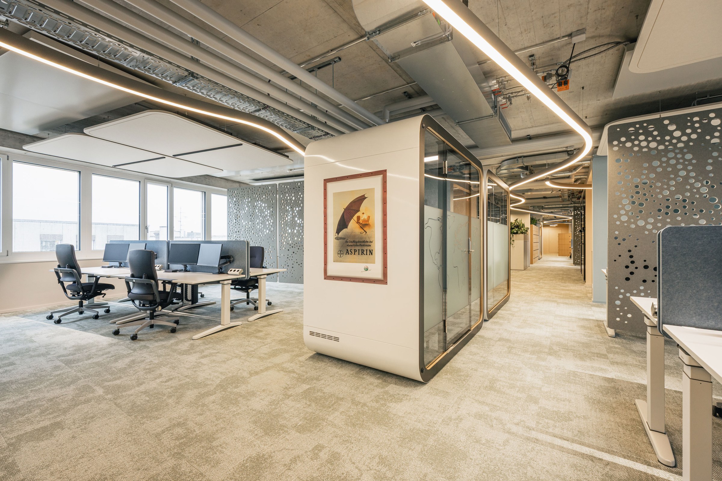 Helles Open-Space-Büro mit Schreibtischen, Telefonbox, dekorativ durchbrochenen Wandpaneelen und sichtbarer Deckeninstallation.
