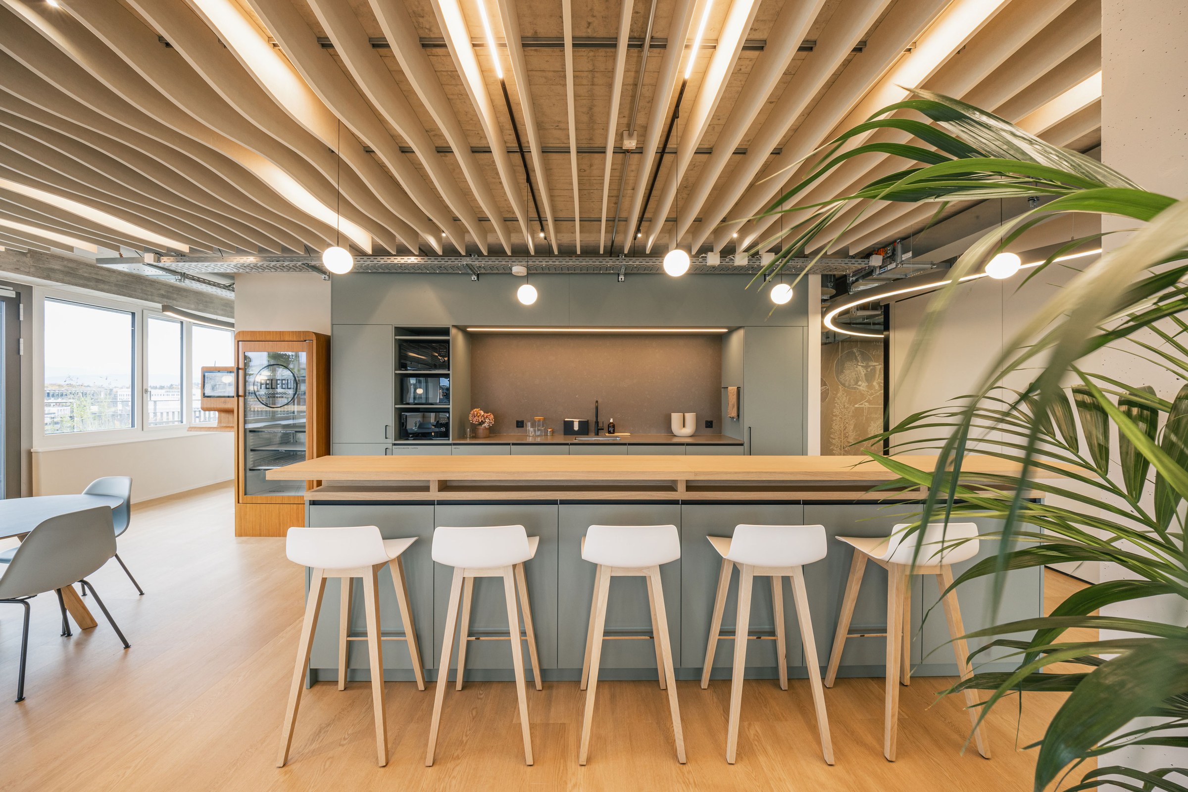 Große, moderne Büroküche mit Holztheke, mehreren weißen Barhockern und einer akzentuierten Deckenbeleuchtung.