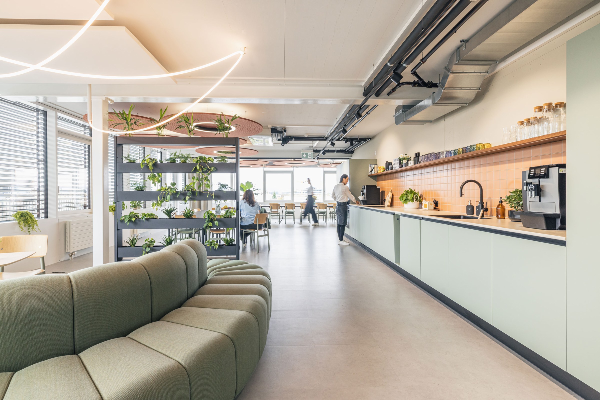 Heller Office-Community-Bereich mit Designsofa, Pflanzenregal und moderner Büroküche.