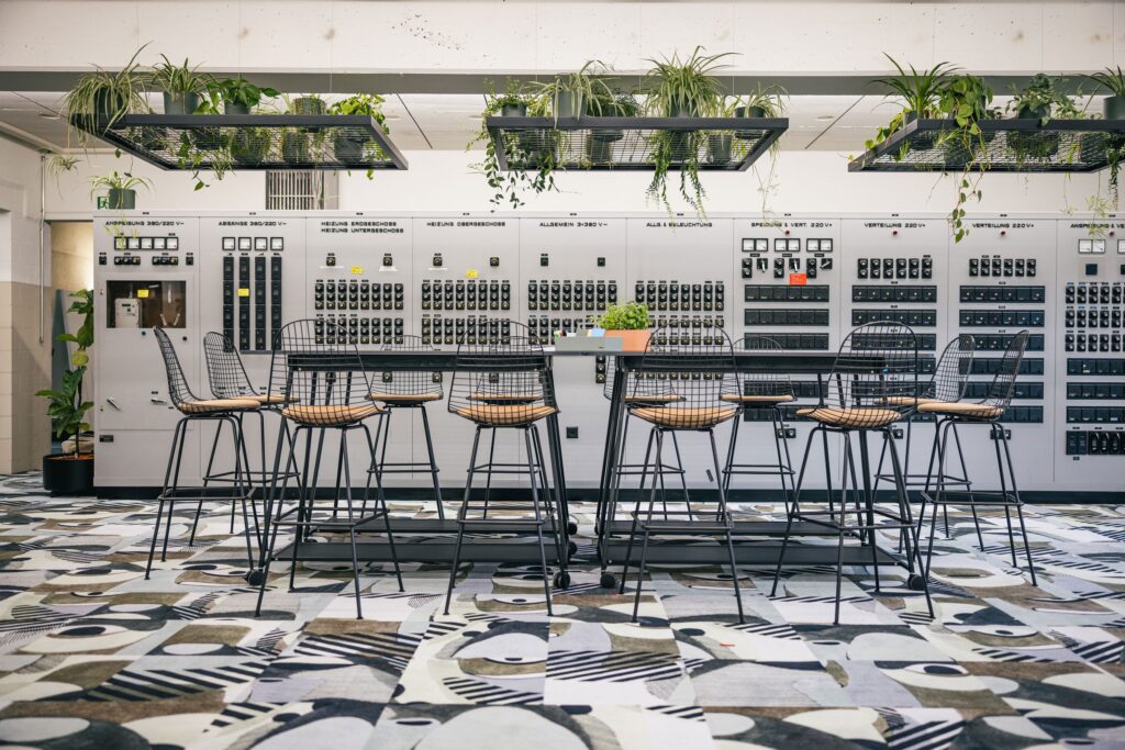 Workshopzone mit großem Tisch, schwarzen Stühlen und farbigem Teppich.