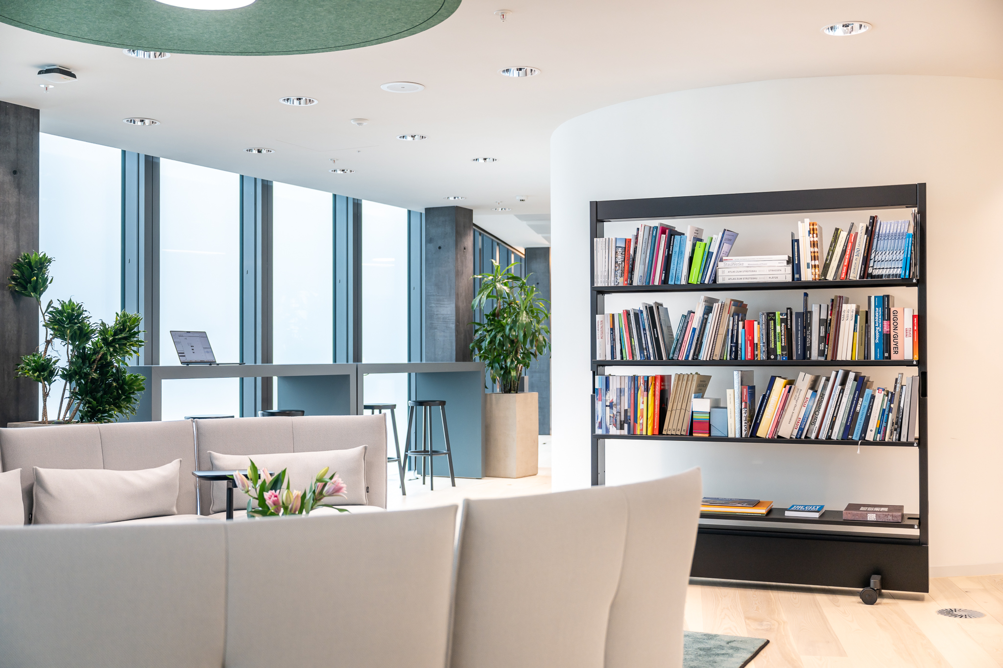 Moderner Loungebereich mit hellen Sofas, großem Bücherregal und hohen Bartischen vor bodentiefen Fenstern.