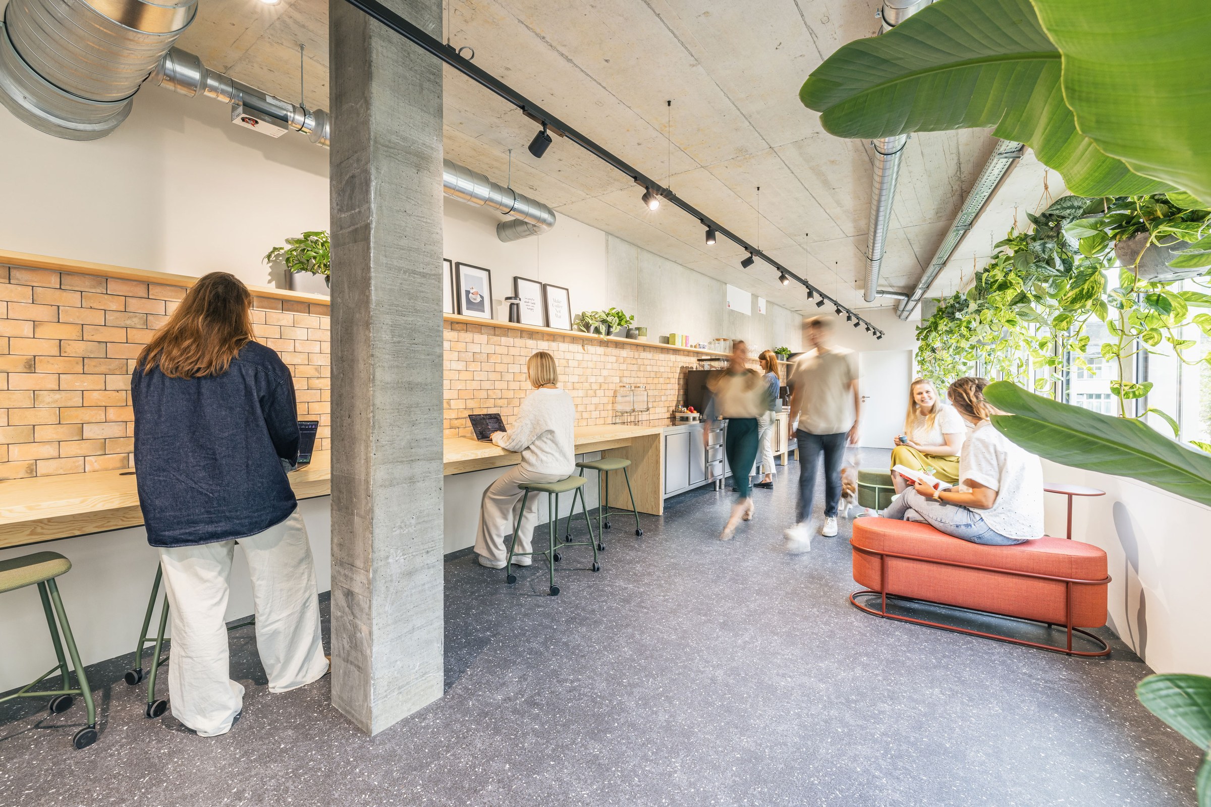 Belebter Bürobereich mit hoher Bar-Arbeitszeile, Backsteinwand und üppigen Pflanzen entlang der Fensterfront.