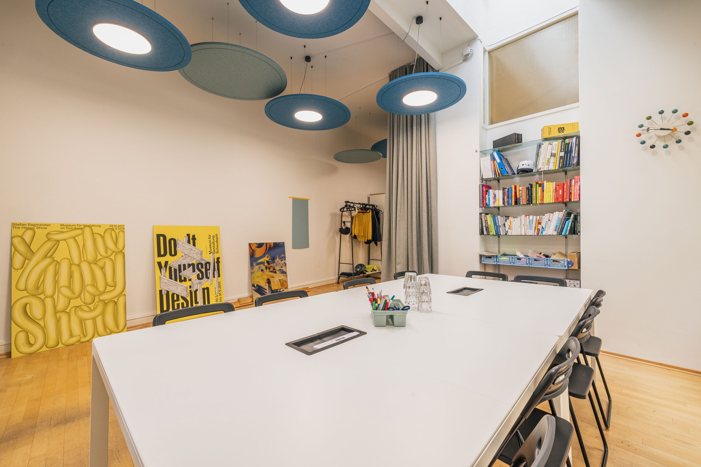 Großer Besprechungsraum mit Akustikdecken, weißen Tischen, farbigen Postern und offenem Bücherregal.