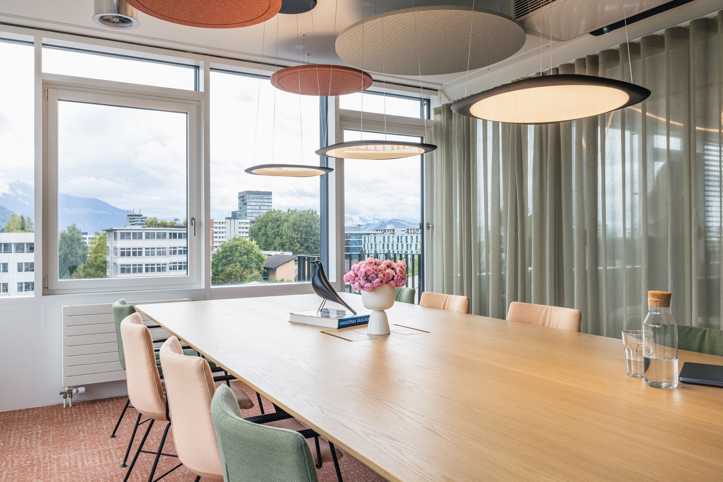 Moderner Konferenzraum mit großem Holztisch, Designerleuchten und Stadtblick durch bodentiefe Fenster.