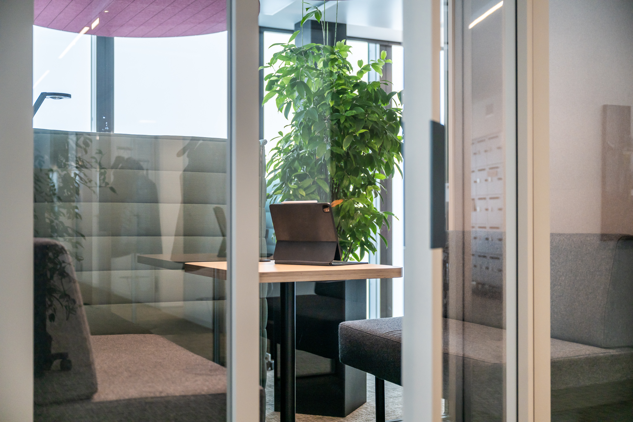 Kleiner Fokusraum mit gepolsterten Booths, Holztisch und Tablet, umgeben von Glaswänden und großer Pflanze.