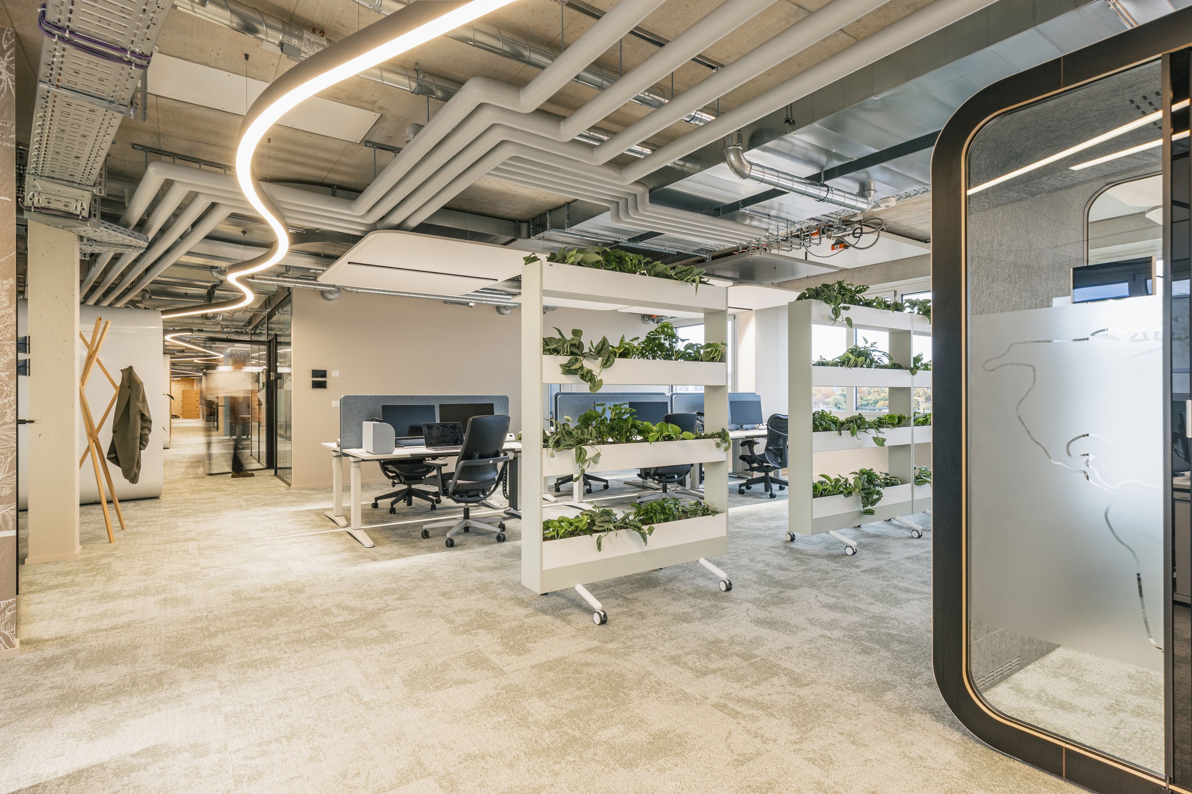 Modernes Open-Space-Büro mit begrünten Raumteilern, mehreren Arbeitsplätzen und geschwungener Deckenbeleuchtung.